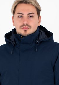 Giacca con cappuccio blu navy caratterizzata da un colletto alto, cordino regolabile e una consistenza liscia, adatta per l'uso all'aperto.