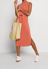 Valaistusoranssi, hihaton mekko, jossa on kiristysvyötärö ja sivurei'ät, yhdistettynä beigen väriseen kassi, jossa on vaaleanpunaista tekstiä. Valkoiset lenkkarit jalassa.