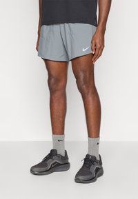 Γκρι αθλητικές βερμούδες Nike με λευκό λογότυπο, συνδυασμένες με μαύρα αθλητικά παπούτσια και γκρι κάλτσες. Οι βερμούδες έχουν ελαφρύ, λείο υλικό.