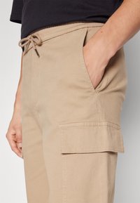 Persoon in beige cargobroek met een trekkoord aan de voorkant en hand in de voorzak, tegen een effen achtergrond.