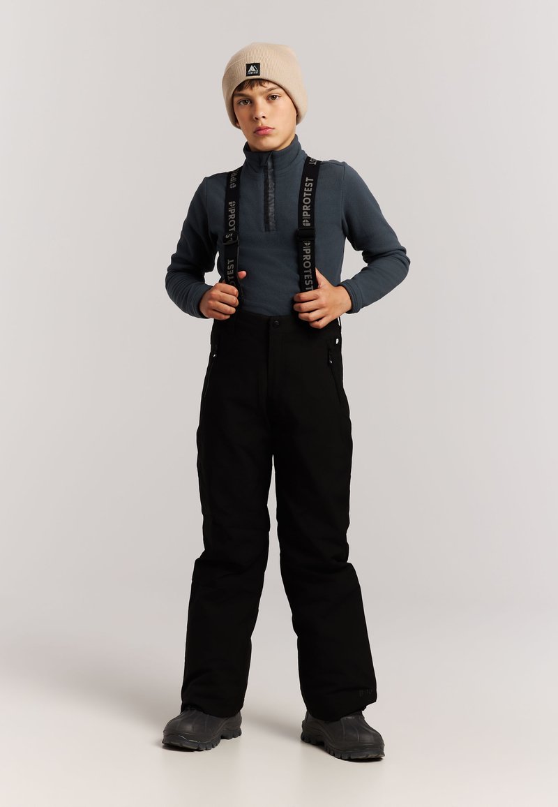 Kind trägt beige Mütze, blaues Langarmshirt, schwarze Schneehose mit Hosenträgern und schwarze Stiefel, steht vor einfarbigem Hintergrund.
