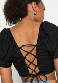 Top preto de manga curta com mangas bufantes, apresentando um design costas cruzadas com atacadores. Tecido suave com corte justo.
