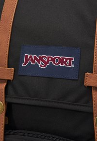 Zaino in tessuto nero con cinturini in ecopelle marrone e una toppa blu con il logo "Jansport" in lettere rosse. Accenti in hardware dorato.