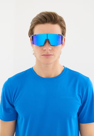 Ung mand iført blå refleks sports solbriller og en matchende blå atletisk t-shirt, set forfra mod en hvid baggrund.