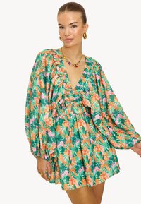 Vestido mini floral con base verde, que presenta patrones tropicales en rosa y naranja. Tiene un escote en V pronunciado, detalles con volantes y mangas abullonadas.