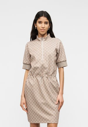 Pikkade tumepruunide juustega naine, kes kannab beežikat, põlveni ulatuvat, mustrilist kleiti lühikeste varrukatega ja esiosas lukuga, seisab ühevärvilise tausta ees.