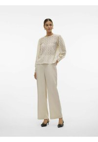 Blouse en dentelle blanc cassé avec un motif géométrique, associée à un pantalon large beige clair et des escarpins à nœud noir. Textures lisses visibles.