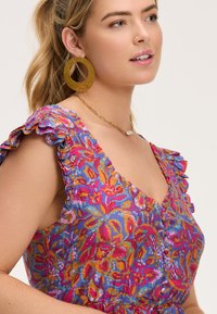 Krāsains ziedu raksts kleitā ar volāniem pie piedurknēm, kas izceļ dzīvīgus rozā, zilu un oranžu toņus. Zelta apaļas auskari un smalka kaklarota.