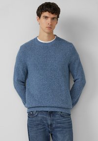 Blauer strukturierter Strickpullover mit Rundhalsausschnitt, gerippten Bündchen und Saum. Wird über einem weißen T-Shirt getragen, kombiniert mit blauen Jeans.