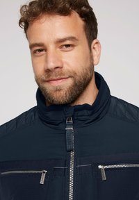 Marineblå jakke med lynlås foran, høj krave og kontrasterende teksturdetailler. Har flere lynlåslommer og et slidstærkt stof.