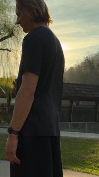 Joven con el cabello hasta los hombros, camisa negra y pantalones cortos, está de pie al aire libre cerca de árboles y un pabellón durante el atardecer.