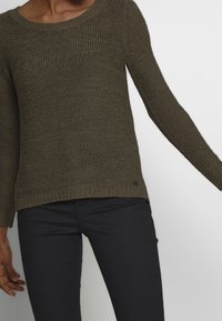 Olivově zelený pletený svetr s kulatým výstřihem, dlouhými rukávy a texturovaným vzorem. Na lemu je malý černý logotyp.