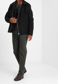 Musta vetoketjullinen takki tummanharmaan paidan päällä, yhdistettynä oliivinvihreisiin housuihin ja ruskeisiin kenkiin. Takissa on kaulus ja se on rento malli.