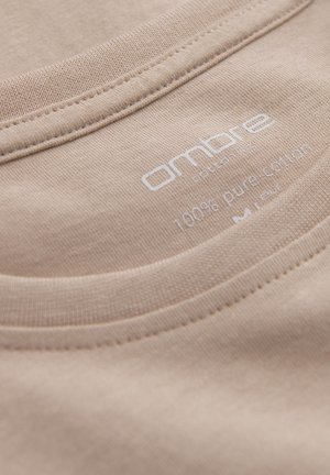 Beige bavlněné tričko s límečkem, hladkou texturou, viditelným švem a potištěnou etiketou uvádějící "100% čistá bavlna."