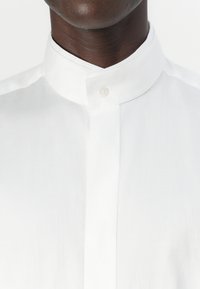 Camicia bianca con pattina e bottoni. Realizzata in tessuto liscio, presenta un collo alla coreana e un design minimalista senza abbellimenti aggiuntivi.
