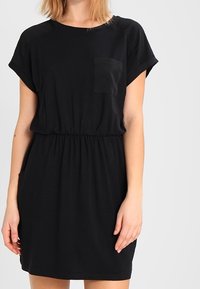 Kobieta w czarnej sukience z krótkim rękawem, z kieszonką na piersi i elastycznym pasem, stojąca na jednolitym tle.