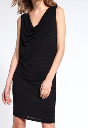 Robe noire sans manches arrivant au genou avec un col drapé portée par une femme debout devant un fond uni.