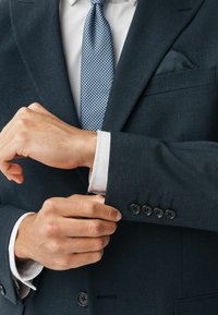 Tmavomodrý textúrovaný oblekový sako s bielou košeľou pod ním. Sako má štyri čierne gombíky na manžetách a vreckovku.
