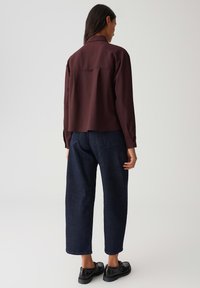 Frau, die mit dem Rücken steht, trägt ein dunkelburgunderfarbenes Langarmshirt, weite dunkelblaue Jeans und schwarze Schuhe vor einem schlicht hellen Hintergrund.