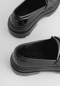 Sorte patenterede loafers med en tyk gummisål, glat overflade og rund form. Indvendigt har de et tekstureret mønster.