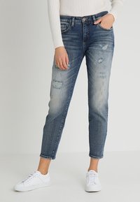 Haalistuneet siniset farkut, joissa on kulutettuja yksityiskohtia ja kapeneva malli, yhdistettynä valkoisiin tennareihin, neutraalia taustaa vasten.