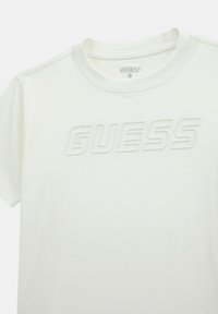 Vit t-shirt med kort ärm som har en upphöjd "GUESS" logotyp i matchande färg, rund halsringning och mjuk bomullstextur.
