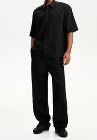 Černá košile s krátkým rukávem na knoflíky s volným střihem, v kombinaci s odpovídajícími volnými černými kalhotami a černou obuví, vyrobená z lehké látky.