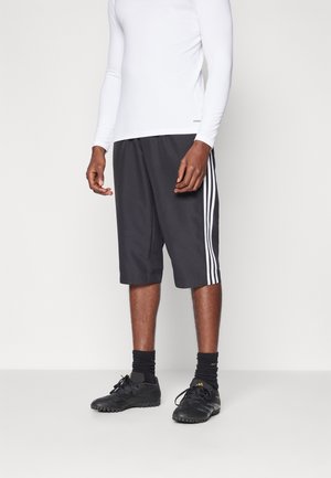 Person trägt schwarze knielange Sportshorts mit weißen Seitenstreifen, schwarze Socken, schwarze Stollenschuhe und ein weißes Langarmshirt.
