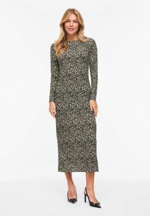 Femme portant une robe midi à manches longues avec un motif zigzag noir et beige, accompagnée de chaussures à talons hauts noires à bride arrière.