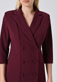 Frau in einem taillierten, dunkelrot-doppelt knöpften Blazer mit drei Viertelärmeln, stehend vor einem schlichten Hintergrund.