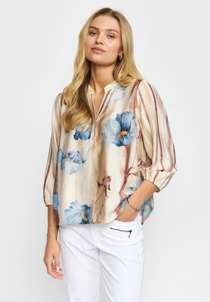 Kvinde med langt blondt hår iført en beige bluse med blå blomsterprint og trekvart-ærmer, kombineret med hvide bukser med lynlåslomme.