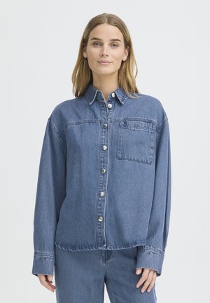 Jeune femme aux cheveux châtain clair portant une chemise en denim bleu boutonnée et un jean assorti, debout devant un fond blanc uni.