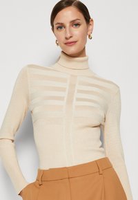 Beige Rollkragenpullover mit strukturierten horizontalen Streifen, geripptem Stoff und einer taillierten Passform, kombiniert mit beigen hochgeschnittenen Hosen.