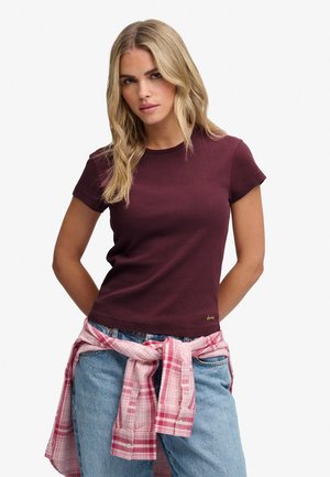 Lyshåret kvinde iført en kortærmet mørklilla t-shirt, blå jeans og en pink ternet skjorte bundet om livet.