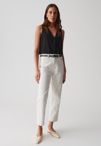 Top sans maniche nero con scollatura a V e bottoni frontali, abbinato a pantaloni bianchi a gamba dritta e una cintura nera con motivo. Scarpe beige con tacco basso.