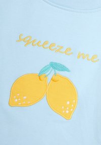 Světle modré tričko z látky s žlutě vyšitými citrony a frází "squeeze me" napsanou žlutým textem nad designem.
