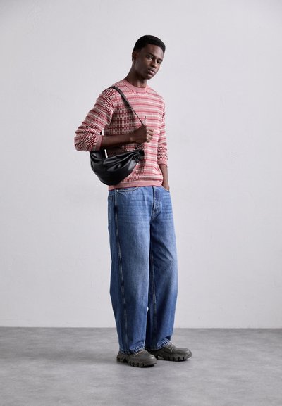 Zwarte schoudertas, rood-wit gestreepte gebreide trui, oversized blauwe spijkerbroek en grijze chunky schoenen, staand tegen een effen achtergrond.