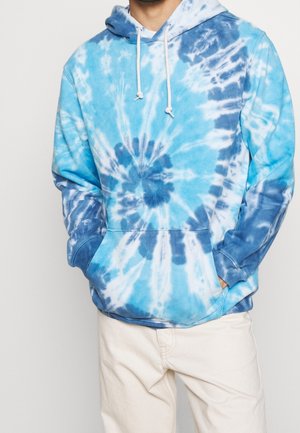 Uomo con felpa a maniche lunghe blu e bianca tie-dye, dotata di tasca frontale e lacci bianchi, abbinata a pantaloni beige chiaro, con le mani nelle tasche.