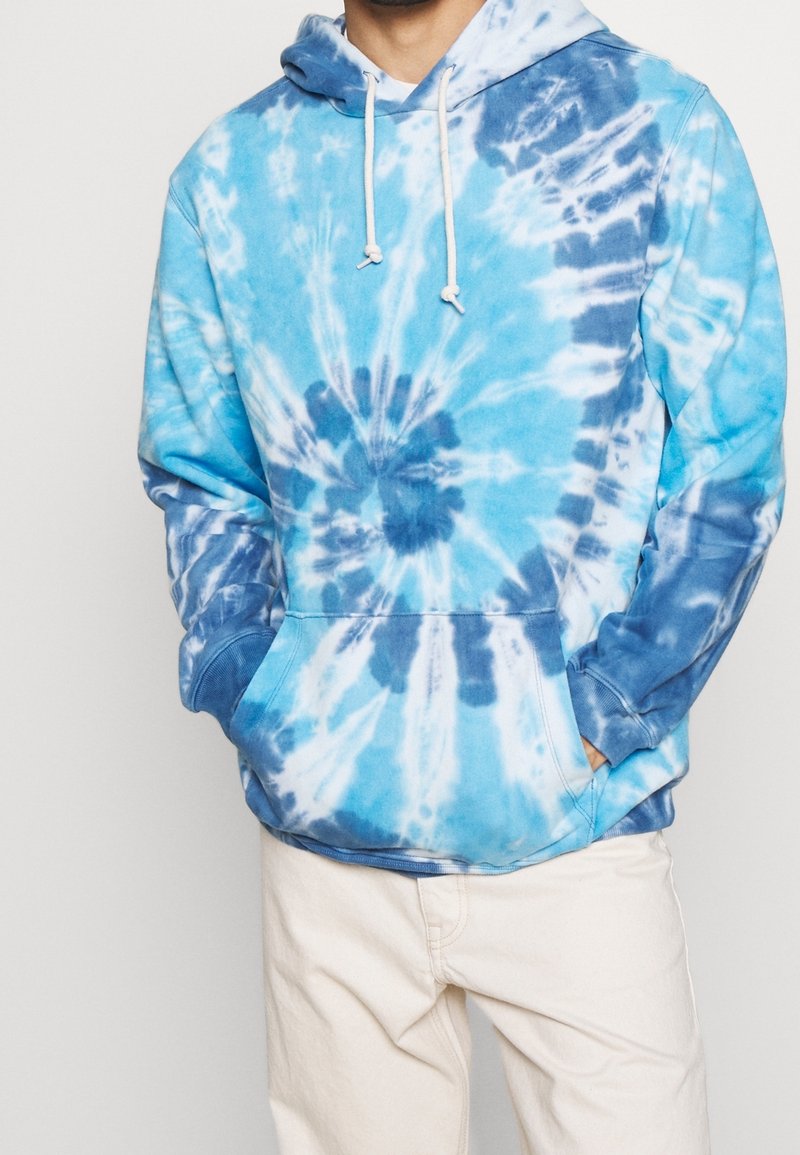 Uomo con felpa a maniche lunghe blu e bianca tie-dye, dotata di tasca frontale e lacci bianchi, abbinata a pantaloni beige chiaro, con le mani nelle tasche.