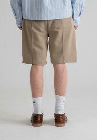 Beige színű pamut rövidnadrág rugalmas derékkal, térd középig érő hosszúságú, hátul zsebbel, fehér bordás zoknival és barna cipővel párosítva.