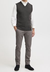 Gilet gris foncé à col en V, en tissu tricoté texturé, porté sur une chemise blanche. Associé à un pantalon gris clair fuselé et des bottines noires.