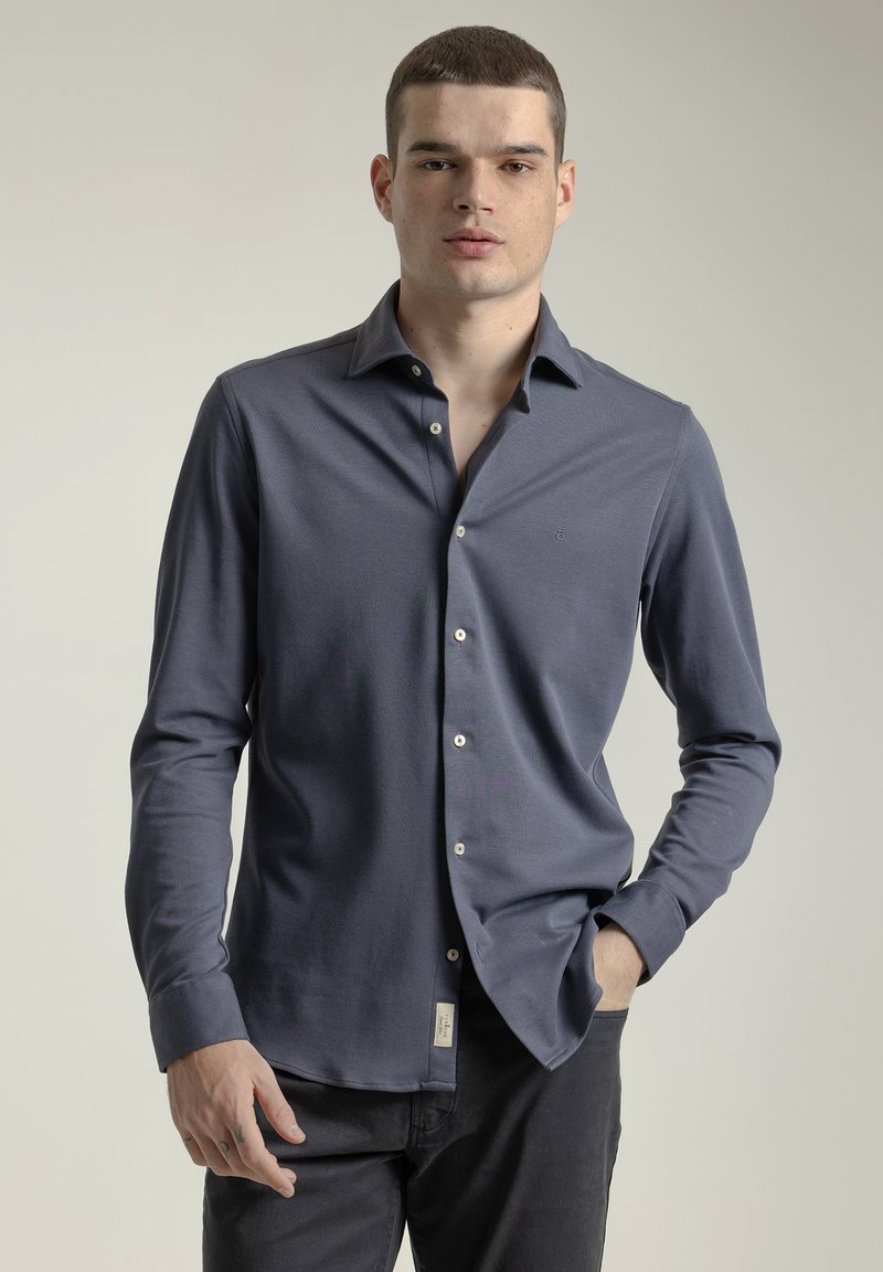 Camisa de manga larga en azul oscuro. Cuenta con cuello clásico, cierre frontal con botones y un logo sutil en el pecho. Textura suave, diseño ajustado.