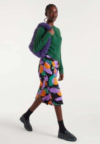 Mujer caminando vestida con un cardigan verde, falda negra con brillantes patrones en naranja y púrpura, calcetines negros, mocasines y cargando una bolsa texturizada púrpura.