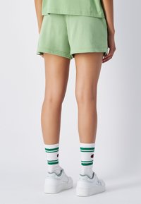 Pantalones cortos de algodón verde claro con dobladillos enrollados, combinados con calcetines blancos que tienen rayas verdes y un acento rojo, usados con zapatillas blancas.