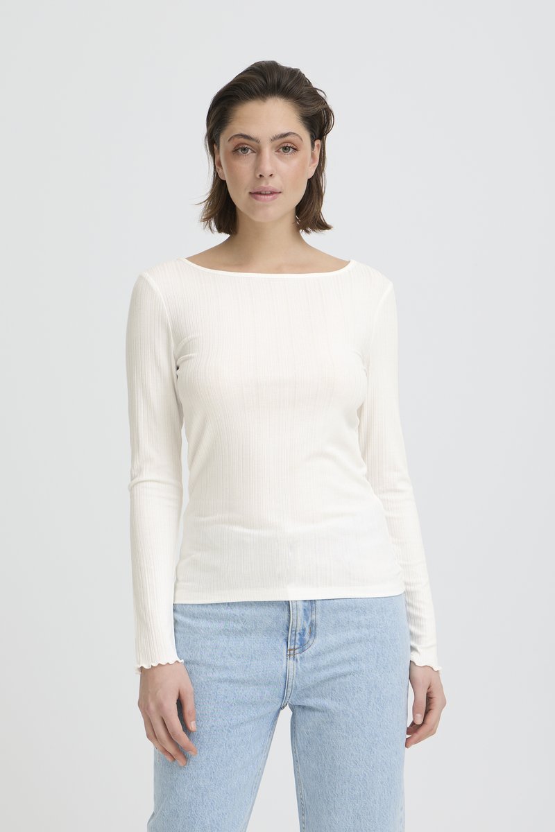 Femme aux cheveux bruns courts portant un haut blanc à manches longues et côtelé ainsi qu'un jean bleu clair, debout devant un fond blanc uni.