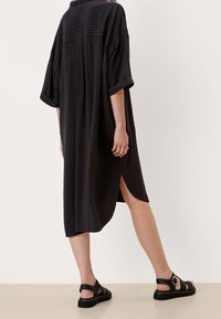 Czarna sukienka w paski o luźnym kroju, z podwiniętymi rękawami i bocznymi rozcięciami, zestawiona z czarnymi sandałami na paski. Gładka faktura i swobodny fason.