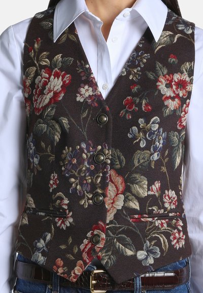 Gilet foncé boutonné avec un motif floral coloré, porté sur une chemise blanche à col, associé à un jean bleu et une ceinture marron.