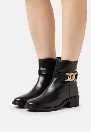 Des bottes noires en cuir jusqu'à la cheville présentent une boucle en métal doré, une texture lisse, un bout arrondi et un petit talon carré. Fermeture éclair sur le côté.