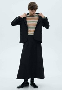 Zwarte op maat gemaakte blazer over een meerkleurige gestreepte trui, gecombineerd met een lange zwarte plissérok en elegante zwarte enkellaarsjes.
