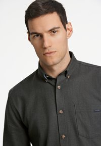 Donkergroene visgraat overhemd met een button-down kraag, voorzien van bruine knopen en een borstzak met een blauw label. Soepele textuur.